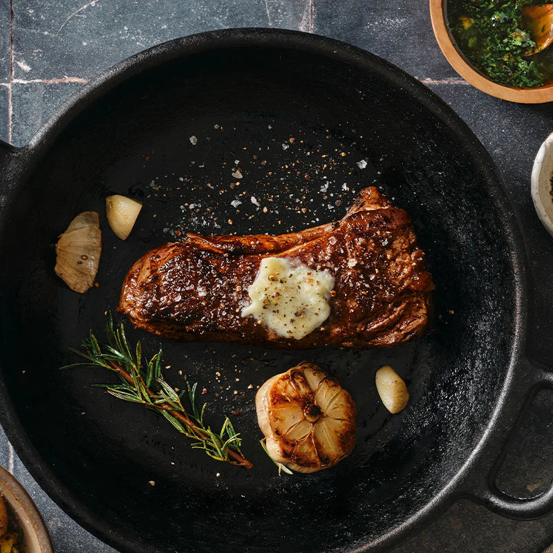 Grilled bison steak with garlic and herbs in a black pan on a dark surface