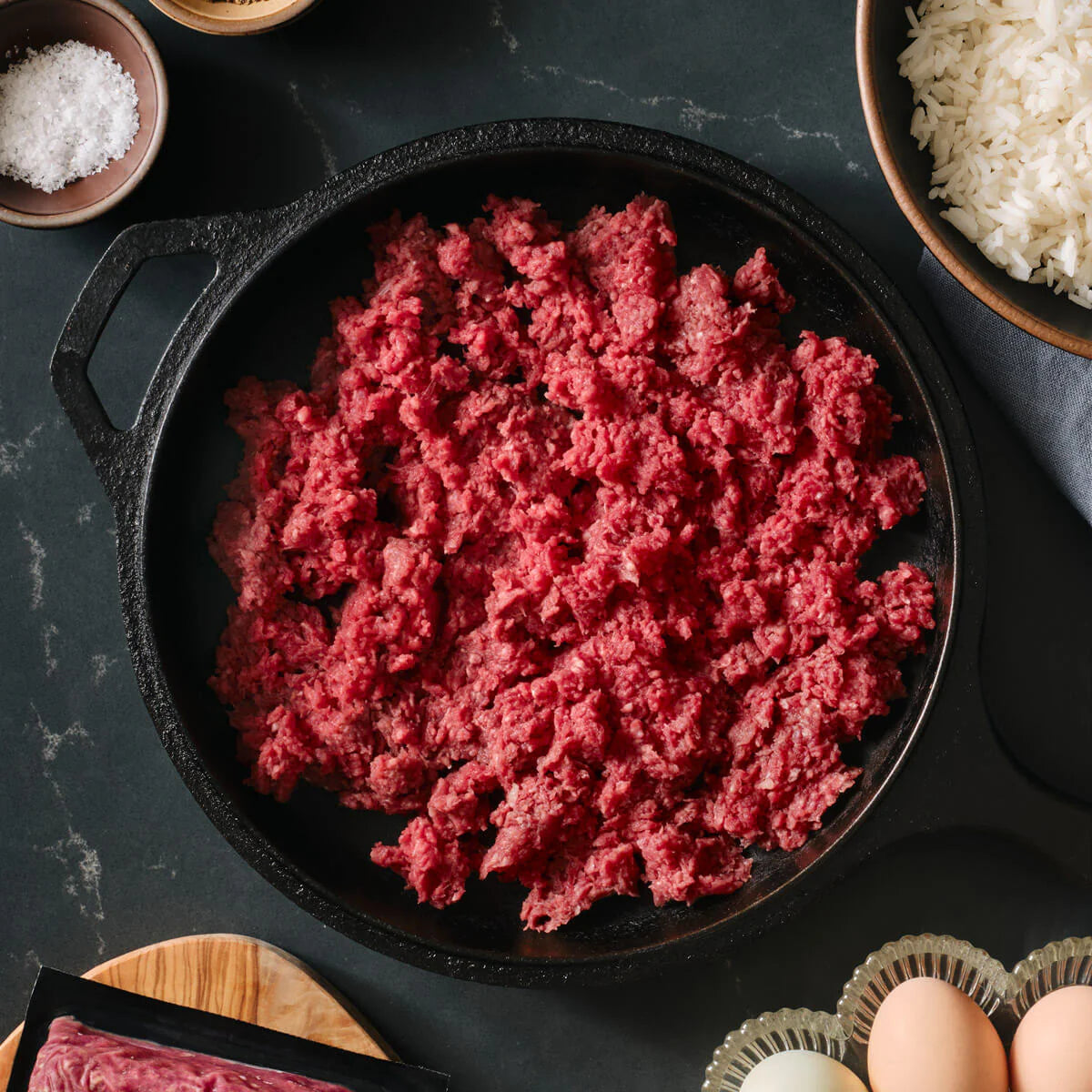 Raw ground beef in a black cast iron skillet on a dark surface with ingredients around.
