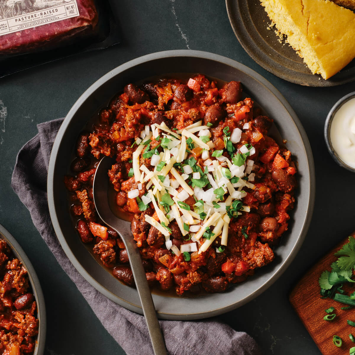 Bowl of venison chili with cheese and green onions on a dark surface with cornbread and a jar of salsa.