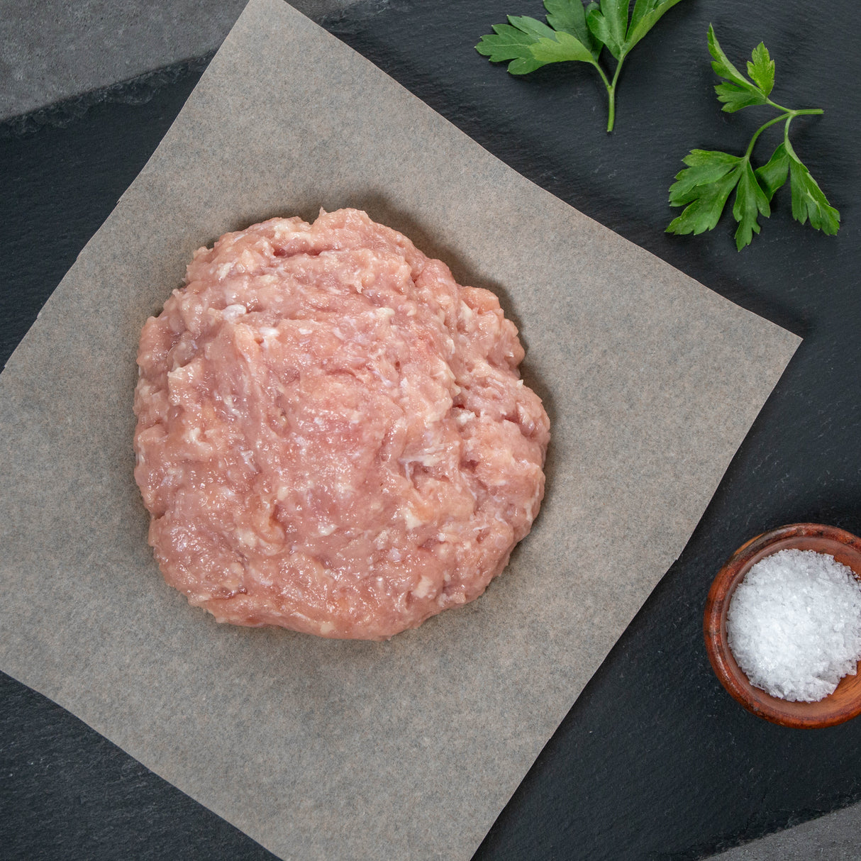 Raw ground chicken patty on brown paper with salt and parsley on a dark surface