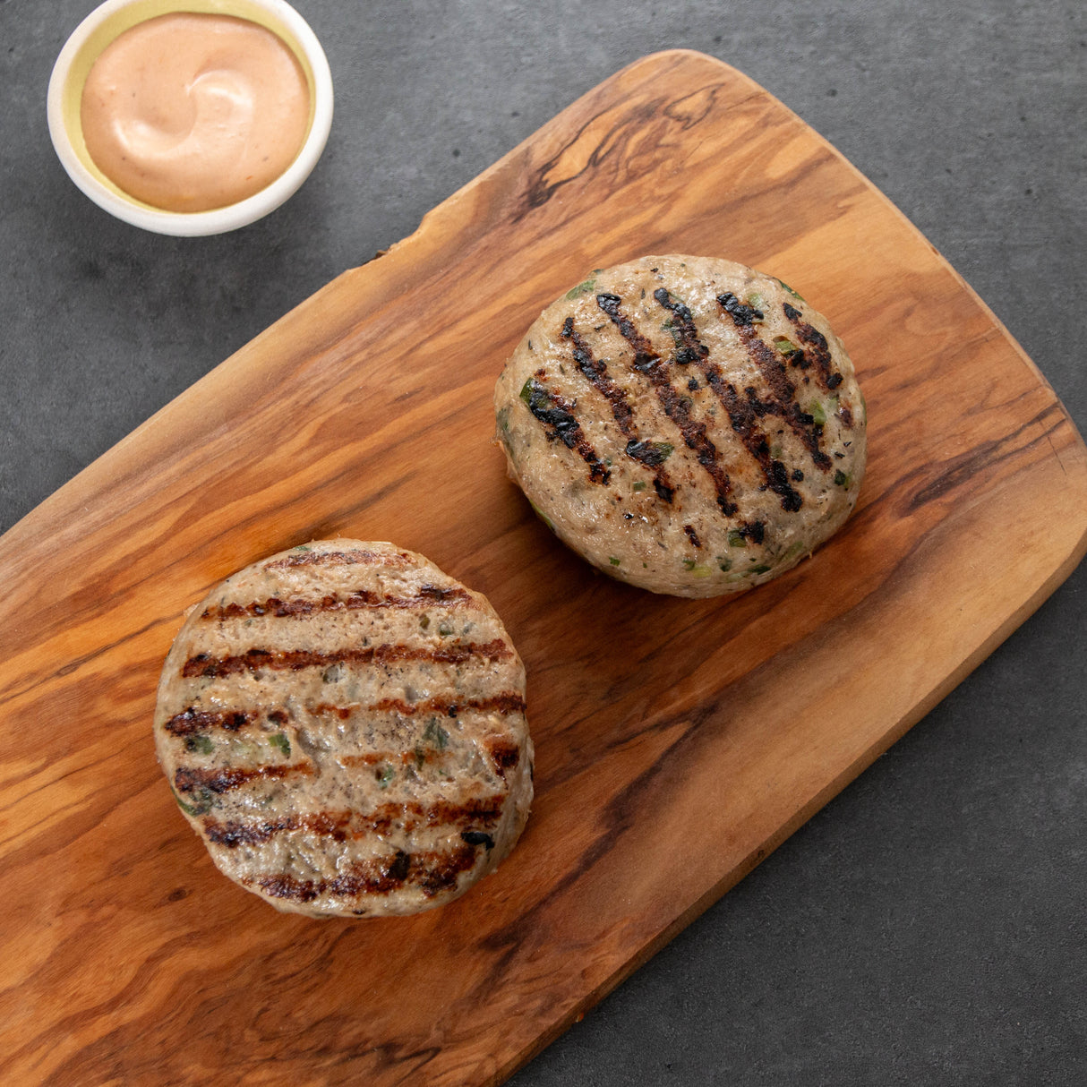 Two grilled turke burger patties on a wooden cutting board with a small bowl of sauce.