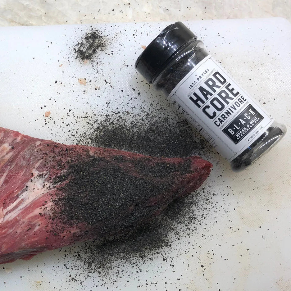 Raw meat with black seasoning on a cutting board next to a bottle of 'Hard Core Carnivore' seasoning.