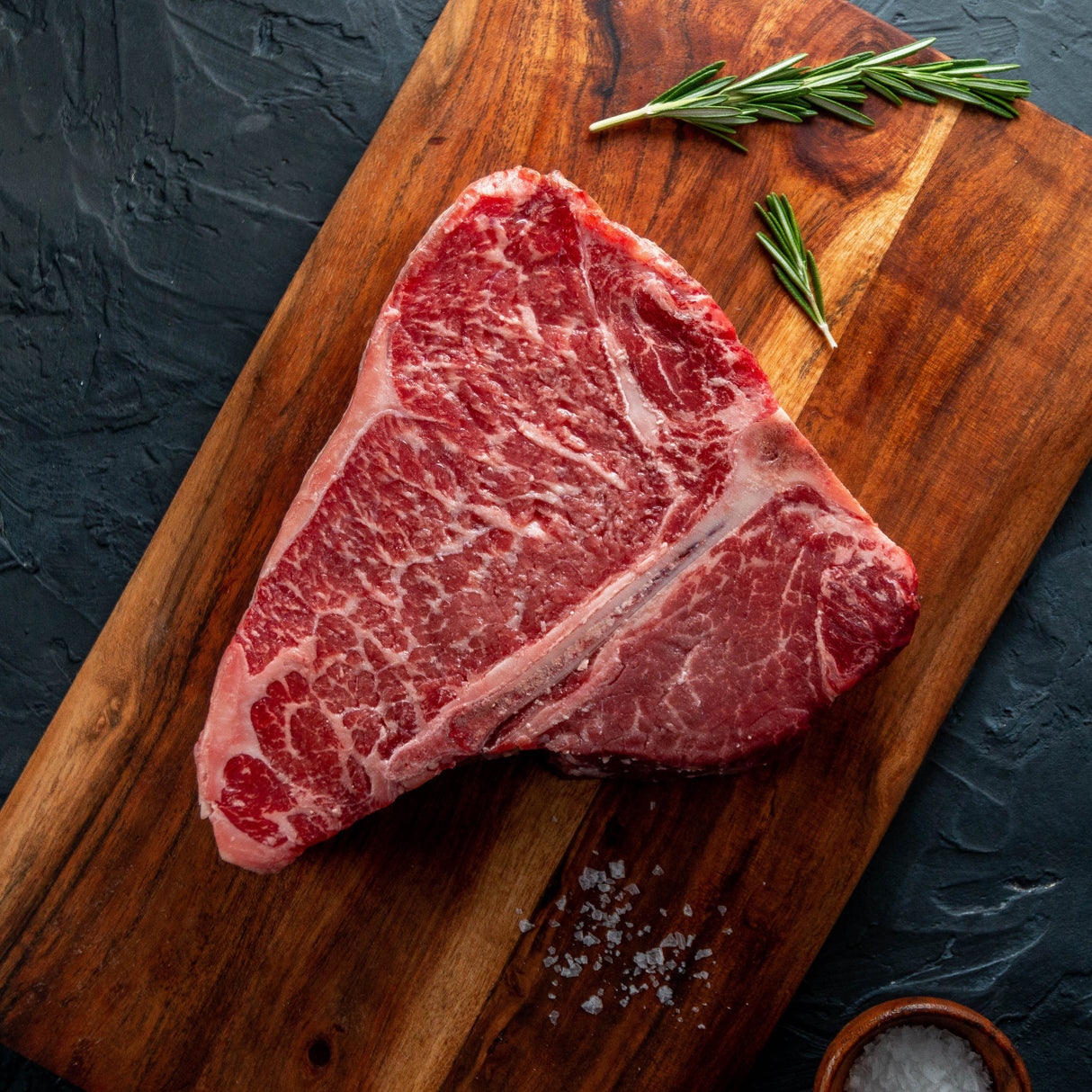 Raw USDA Prime porterhouse steak on a wooden cutting board with rosemary and salt, against a dark background