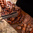 Wooden cutting board with various sausages and a pair of tongs, outdoors.
