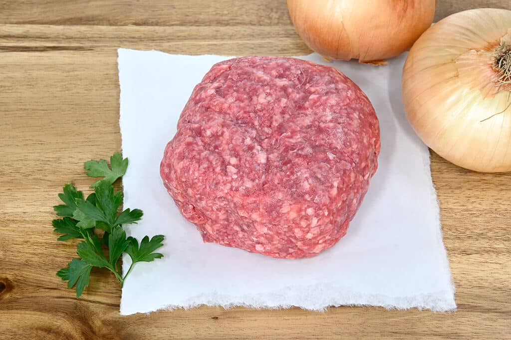 Raw ground meat patty on a wooden cutting board with onions and parsley.
