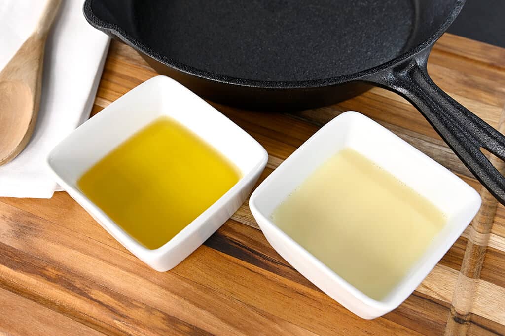 Two small white bowls with oil and another liquid on a wooden surface next to a black cast iron skillet.