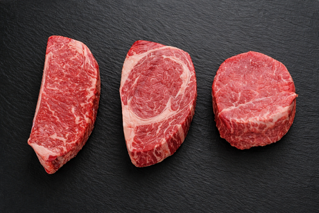 Three SignaturePRIME cuts side by side — NY Strip, Ribeye, and Filet Mignon — on a dark slate surface