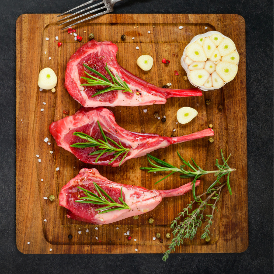 raw lamb rib chops topped with rosemary and a cut garlic head in half on a wooden cutting board