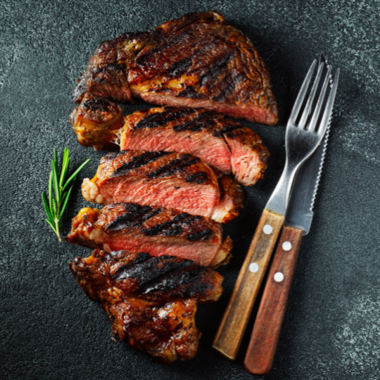 grilled boneless ribeye steak sliced on a slate board with a fork and knife next to it.